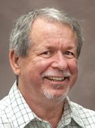 A man with gray hair and a beard is smiling at the camera. He is wearing a light-colored, checkered shirt and standing against a plain, neutral background.